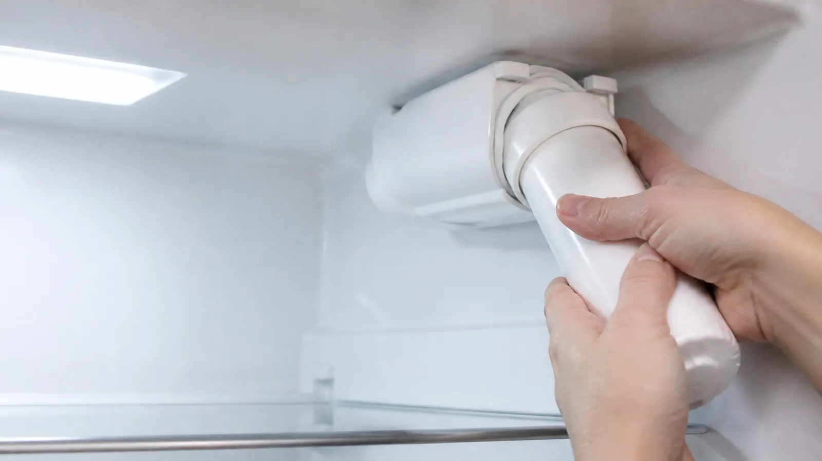 Two hands twisting a plain white water filter cartridge into its housing at the top of a refrigerator's fresh-food compartment