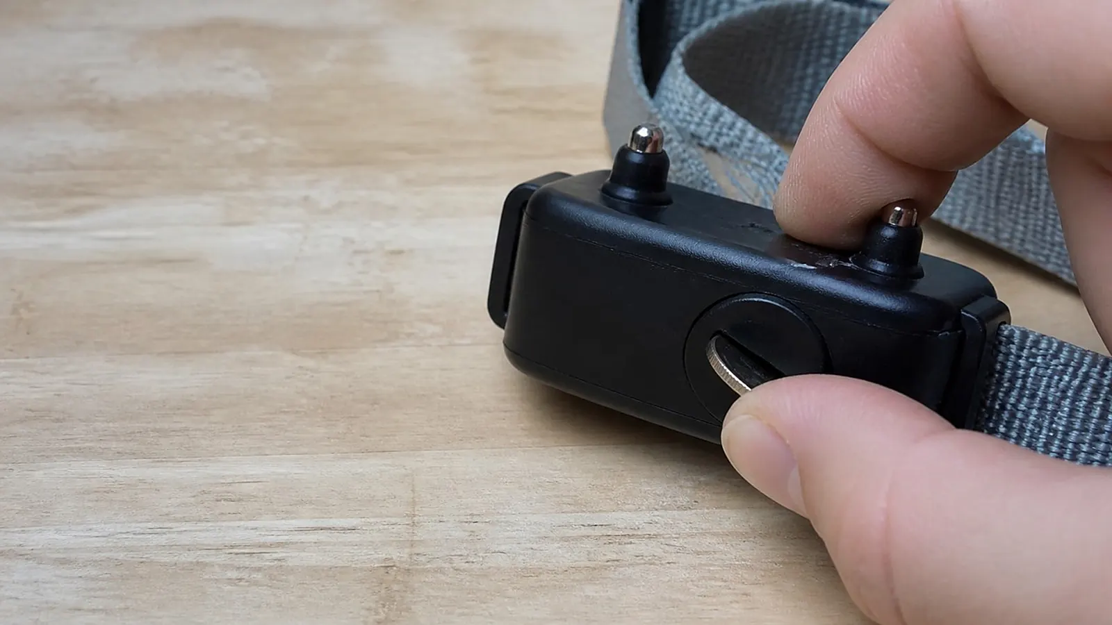 Fingers turning a coin in the battery cap on the underside of a black collar receiver with a gray strap, on a light wood bench