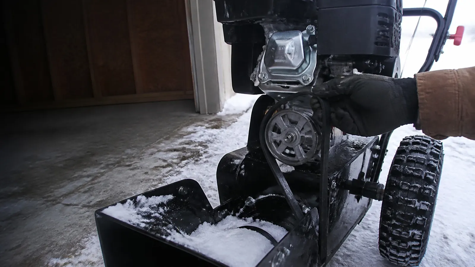 Gloved hand checking the pulley and belt area on the front of a snow blower engine just inside an open garage door