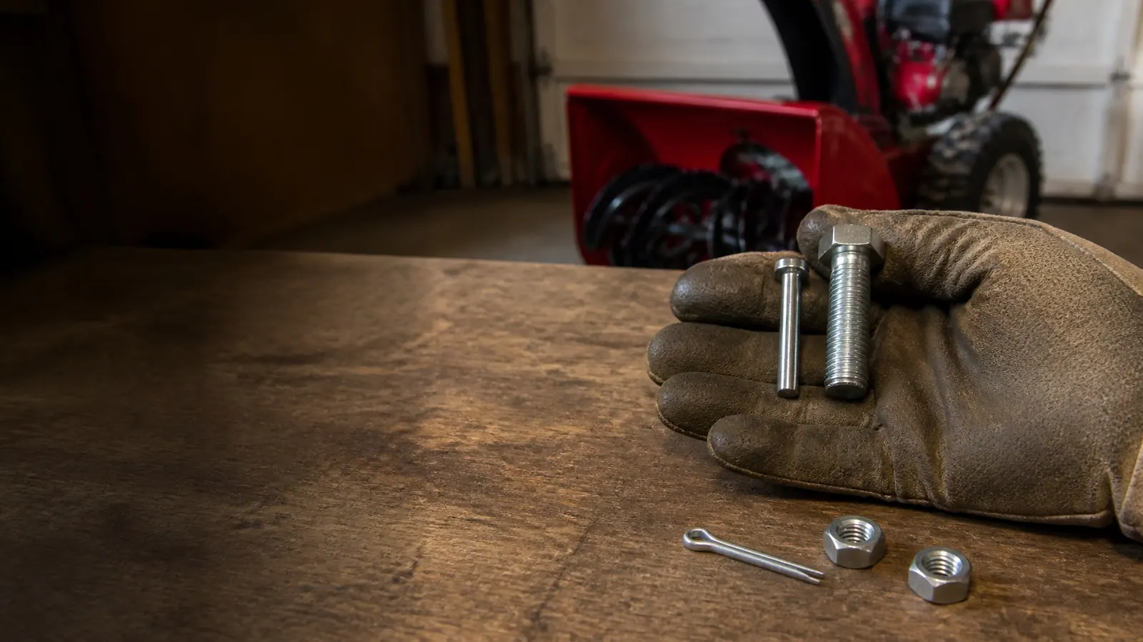 Gloved hand on a garage workbench holding a slim shear pin next to a thicker hex bolt, with nuts and a cotter pin nearby and a snow blower behind