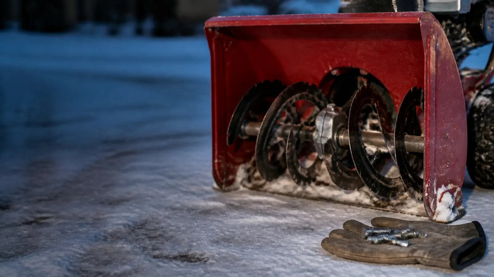 Front of a red two-stage snow blower on an icy driveway at dusk, auger spirals on the shaft, with a work glove holding spare shear bolts in the snow