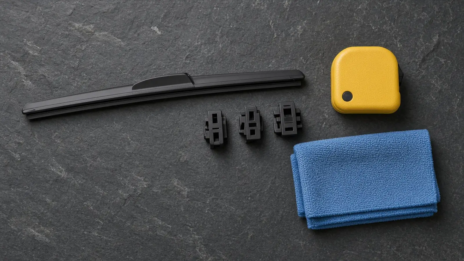 Beam windshield wiper blade, three black plastic arm adapter clips, a tape measure and a folded blue microfiber cloth on a slate workbench