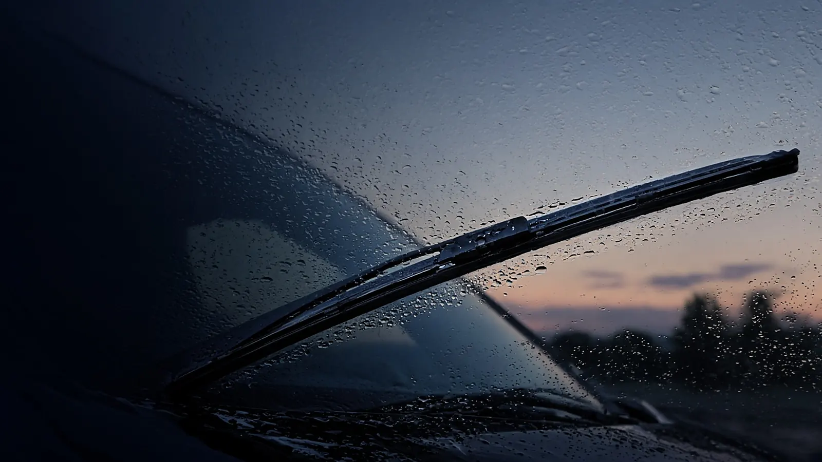 Black beam windshield wiper blade sweeping rain droplets across a car windshield at dusk